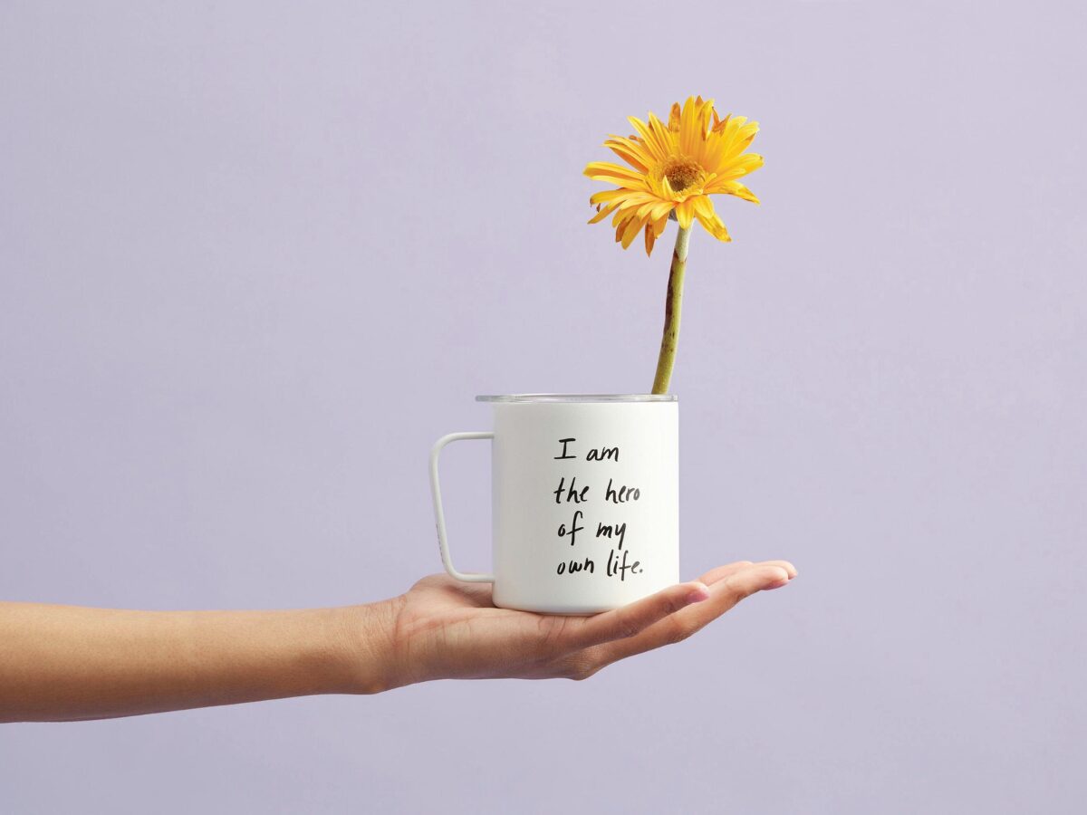 White mug with words “I am the hero of my own life,” holding yellow daisy—an everyday visual reminder of affirmations for real life.