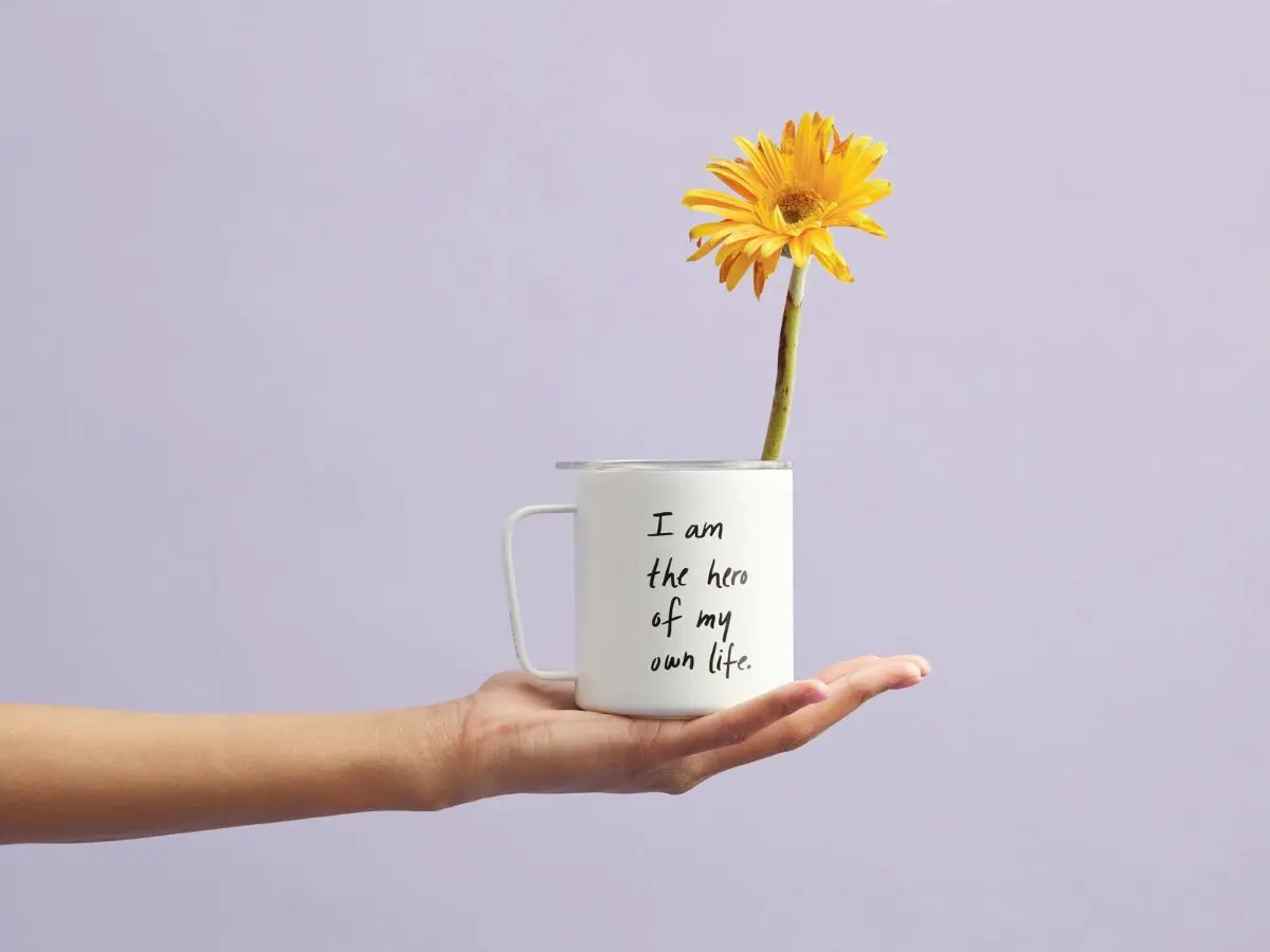 White mug with words “I am the hero of my own life,” holding yellow daisy—an everyday visual reminder of affirmations for real life.